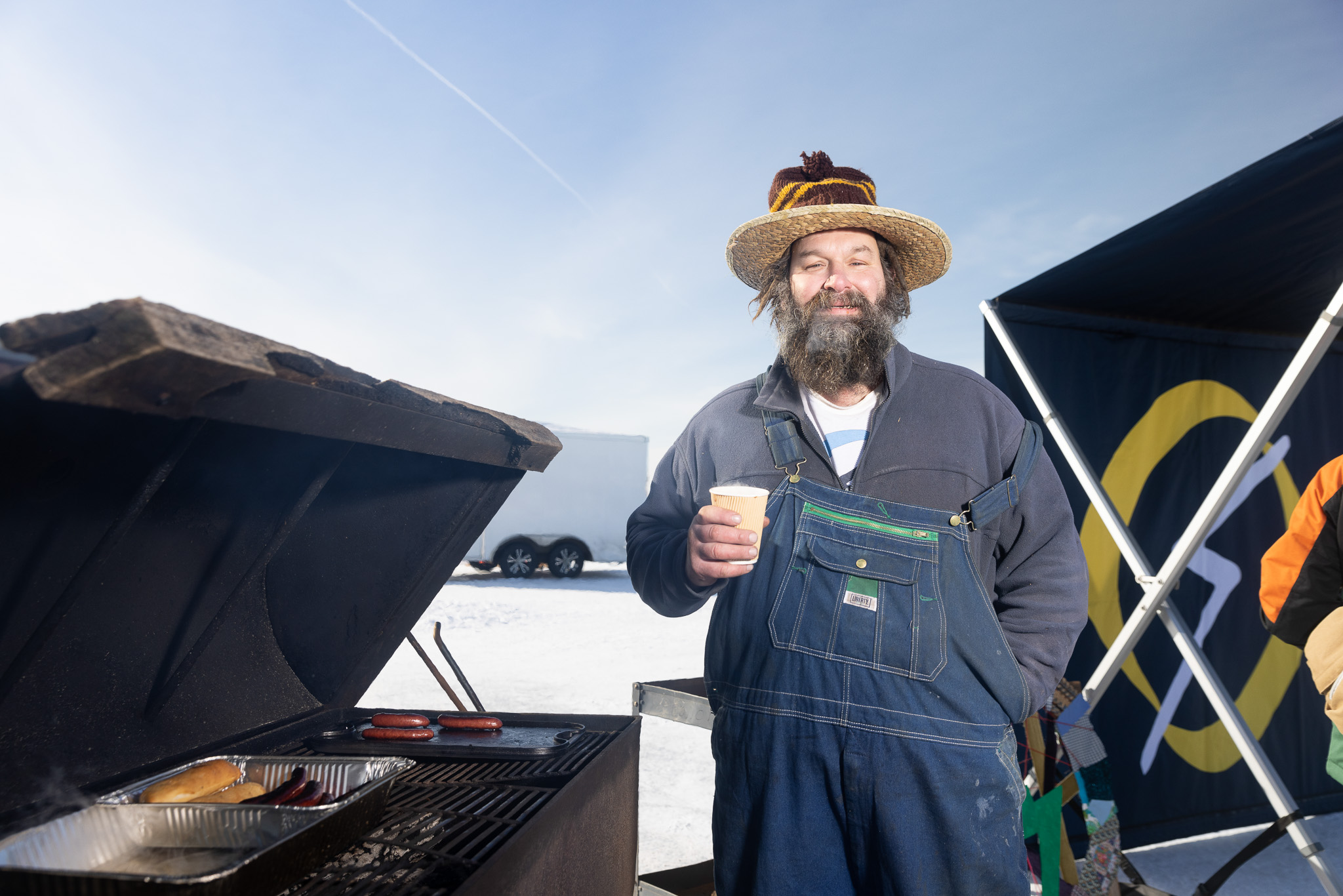 A man in overalls grills hot dogs at an ice fishing tournament.