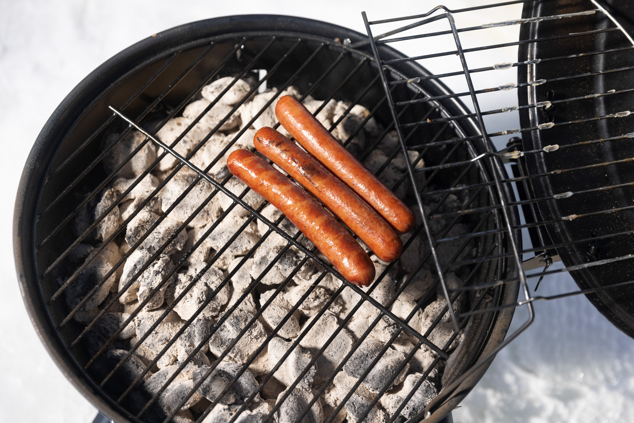 A small charcoal grill with three hot dogs sizzling on the ice.