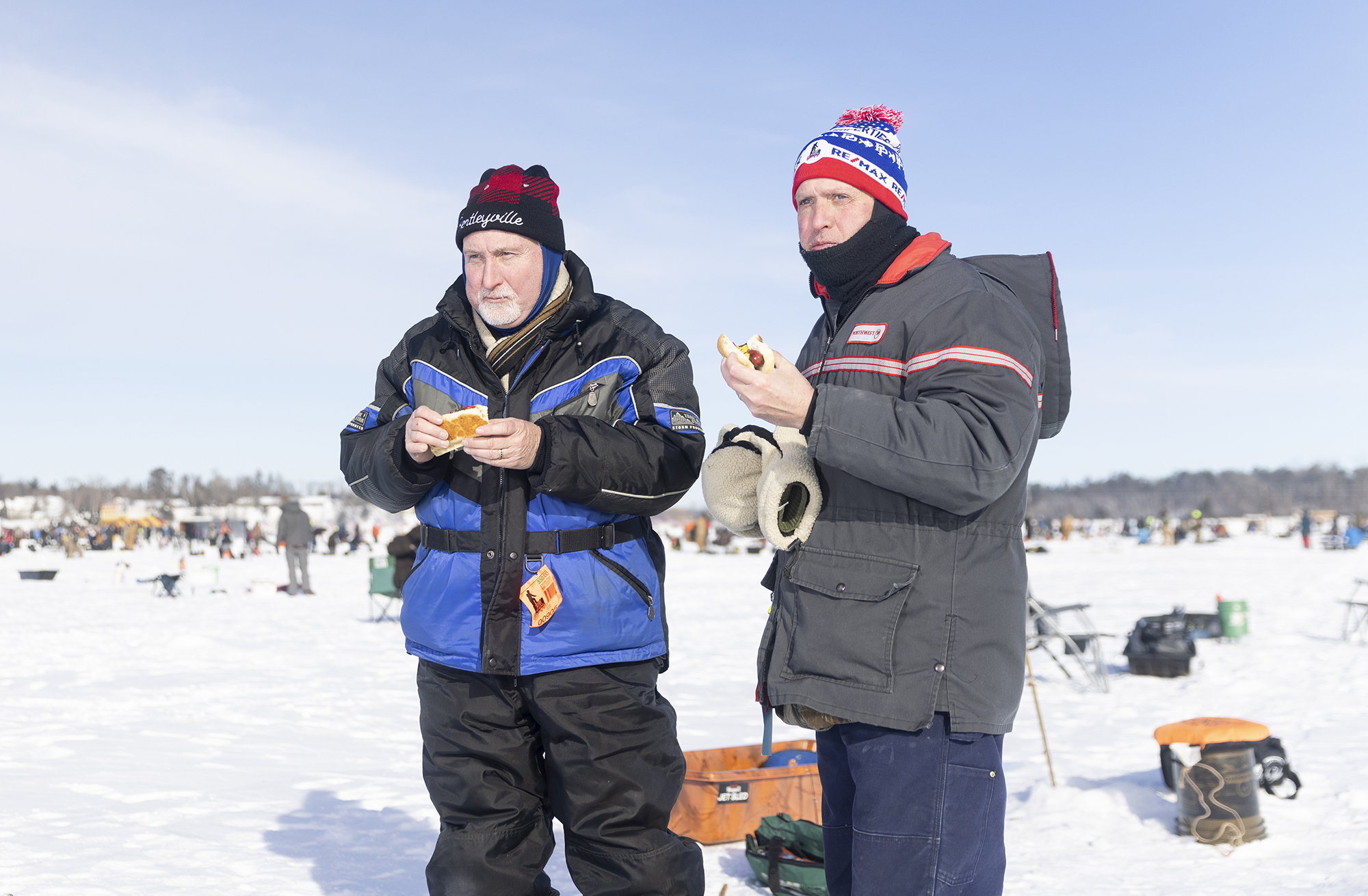 Two men dressed in coats and hats eat hot dogs on the ice.