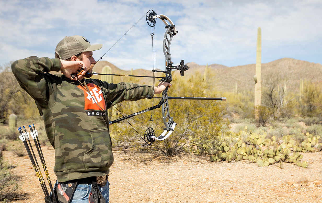 A man in a camo hoodie shoots a PSE bow.