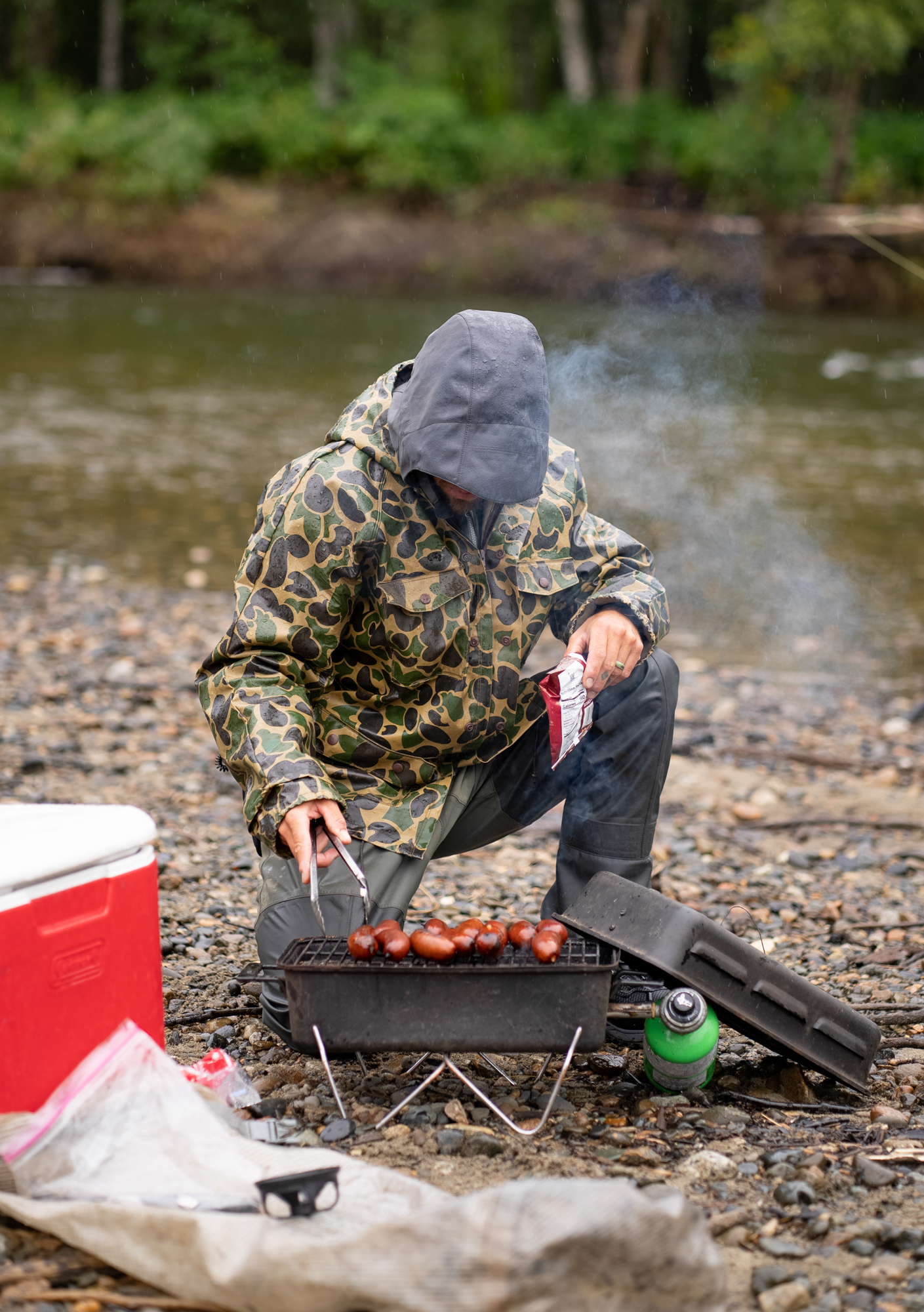 A man in a camo rain jacket grills hotdogs on the bank of a river in Alaska.