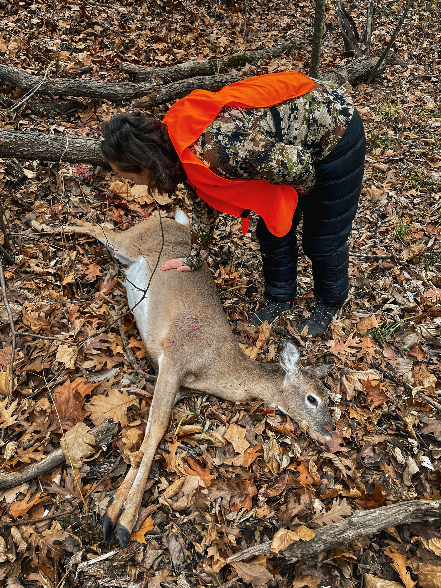 A hunter in a blaze orange vest rests her hand on the side of a whitetail doe.