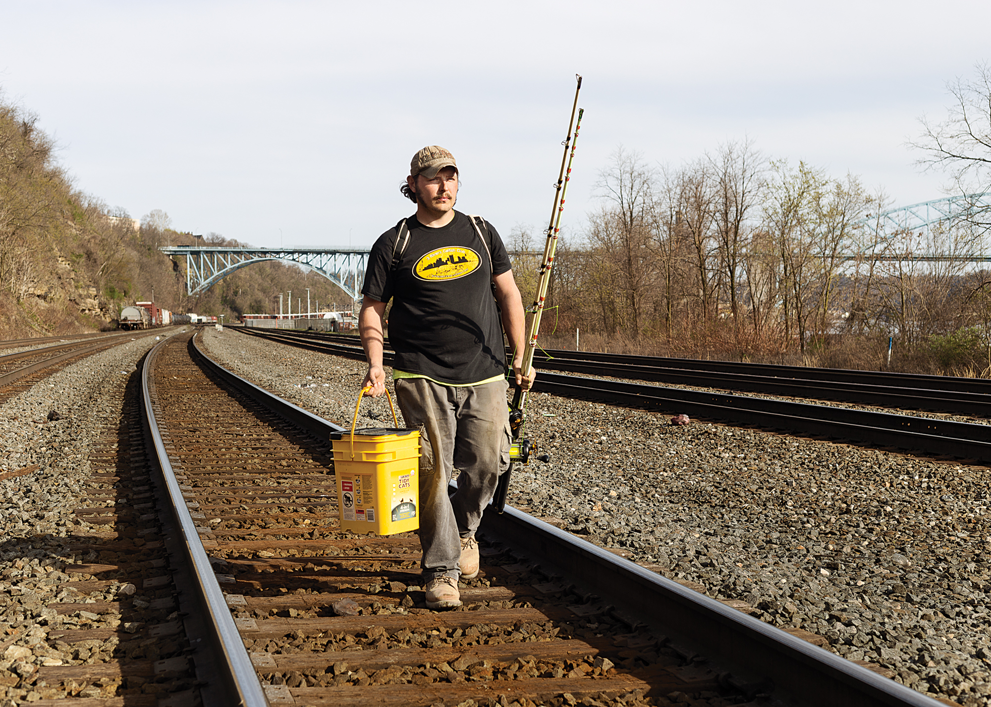 A man with a bucket and fishing rods walks on railroad tracks.