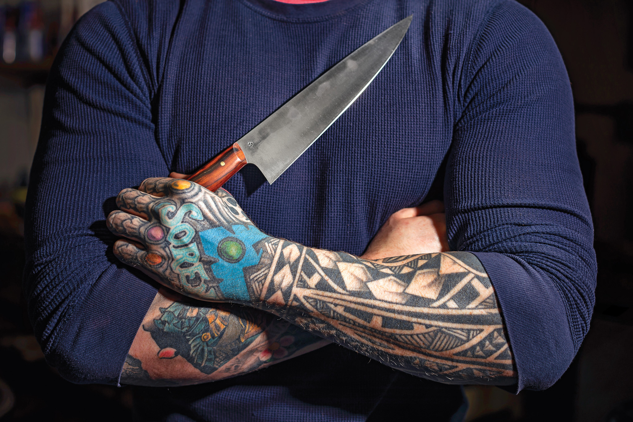 A tattooed knifemaker in Portland holds a big chef's knife across his chest.