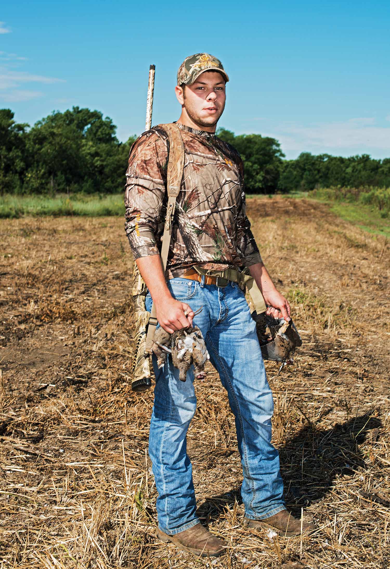 Hunter with shotgun strap over shoulder holds several doves and stands in cut field.