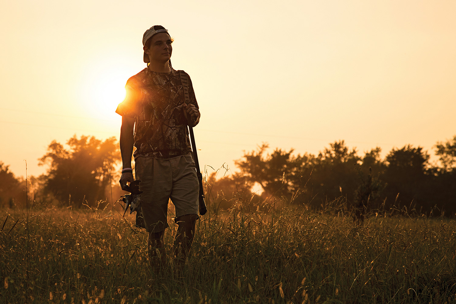 young hunter carrying dove and gun walks through field and sun sets over his shoulder