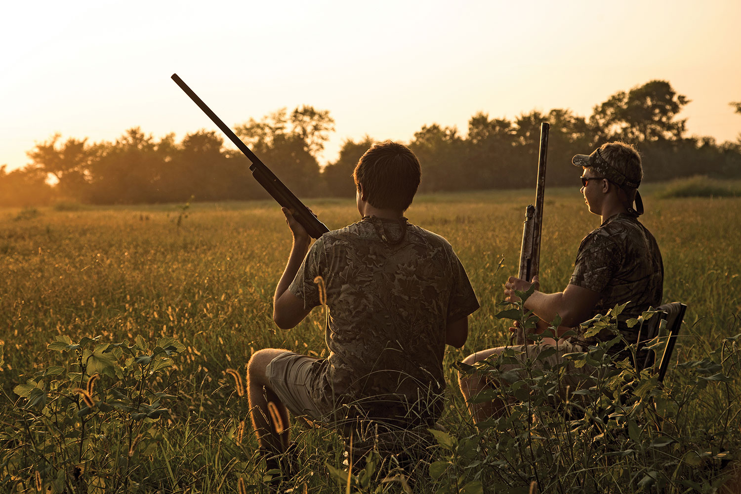 two young dove hunters with shotguns sit in field as sun sets in distance behind trees