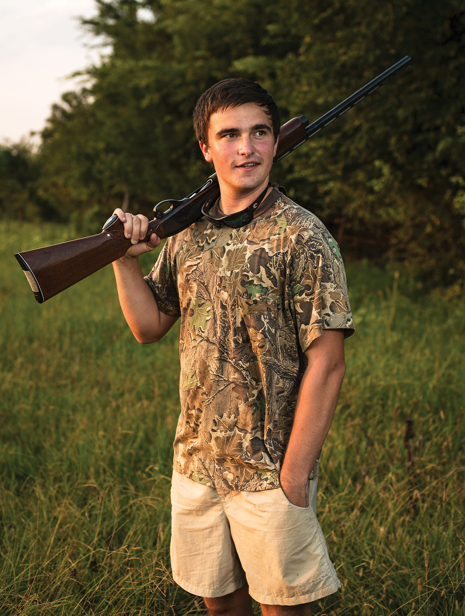 Young dove hunter in camo t-shirt stands in meadow with gun over his shoulder.