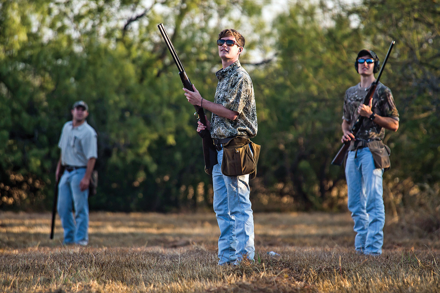 Three young dove hunters with shotguns stand in open field looking for birds.
