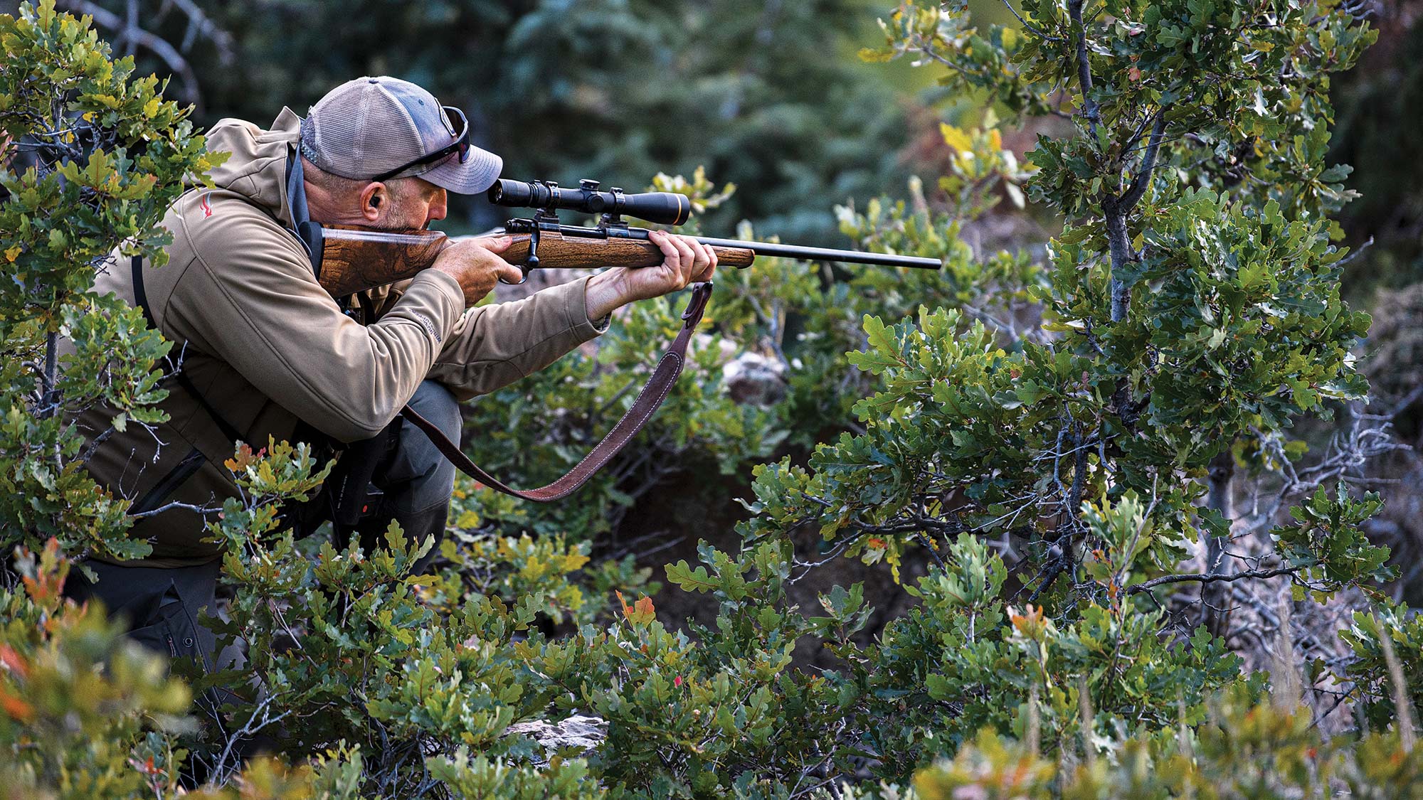 Hunter peers through riflescope and crouches in low brush.