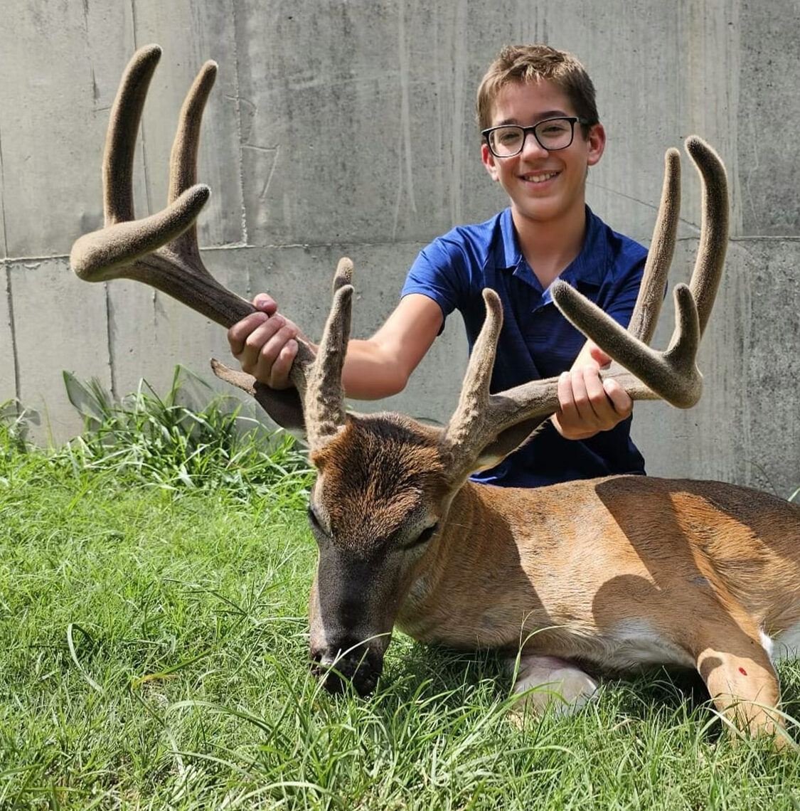 A 10-year-old kid with glasses holding the antlers of a big typical velvet buck.