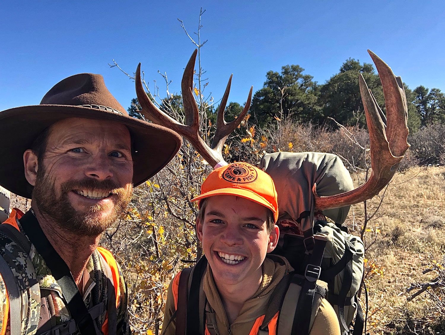 two hunters pose for selfie, one carrying a pack with large mule deer antlers