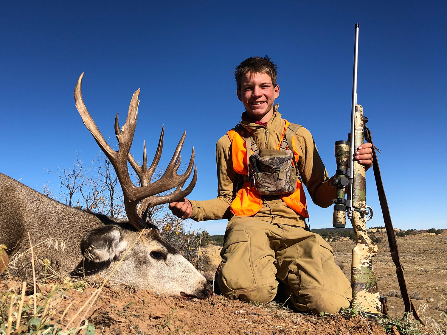 smiling young hunter holding rifle kneels next to downed mule deer