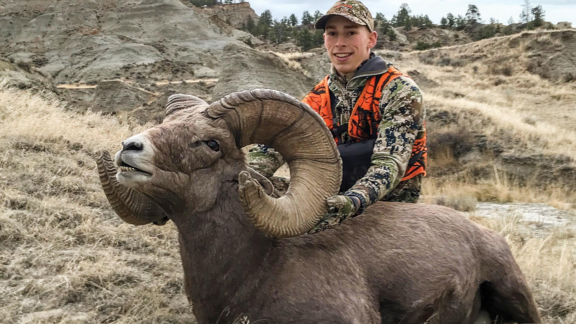 hunter poses with record-book bighorn ram in rocky, grassy area