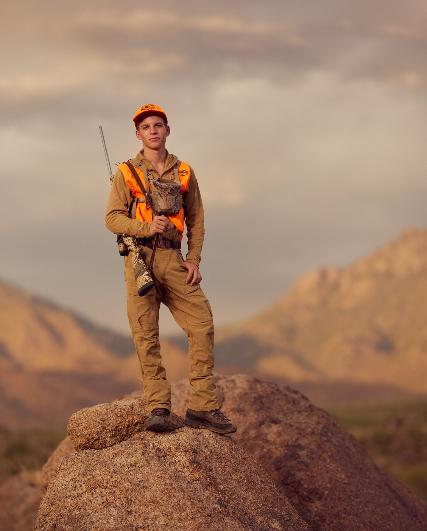 A hunter in an orange vest with a rifle stands on a rock.