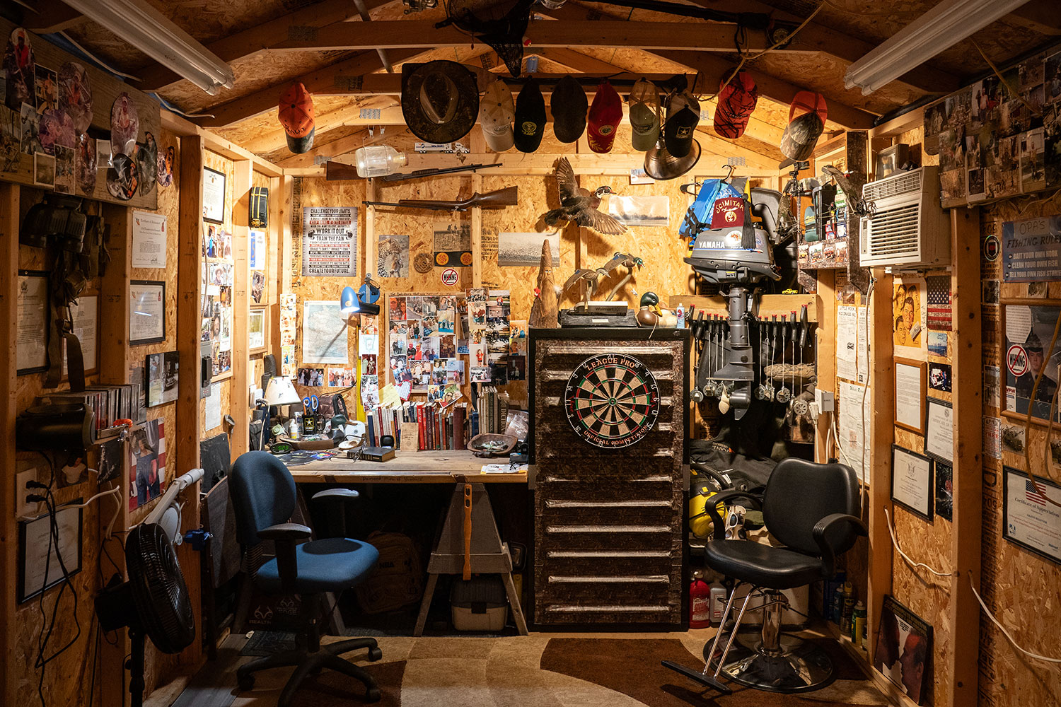 interior of shed-office has many photos, hats, and other wall decorations, plus dartboard, chairs, boat motor, and more