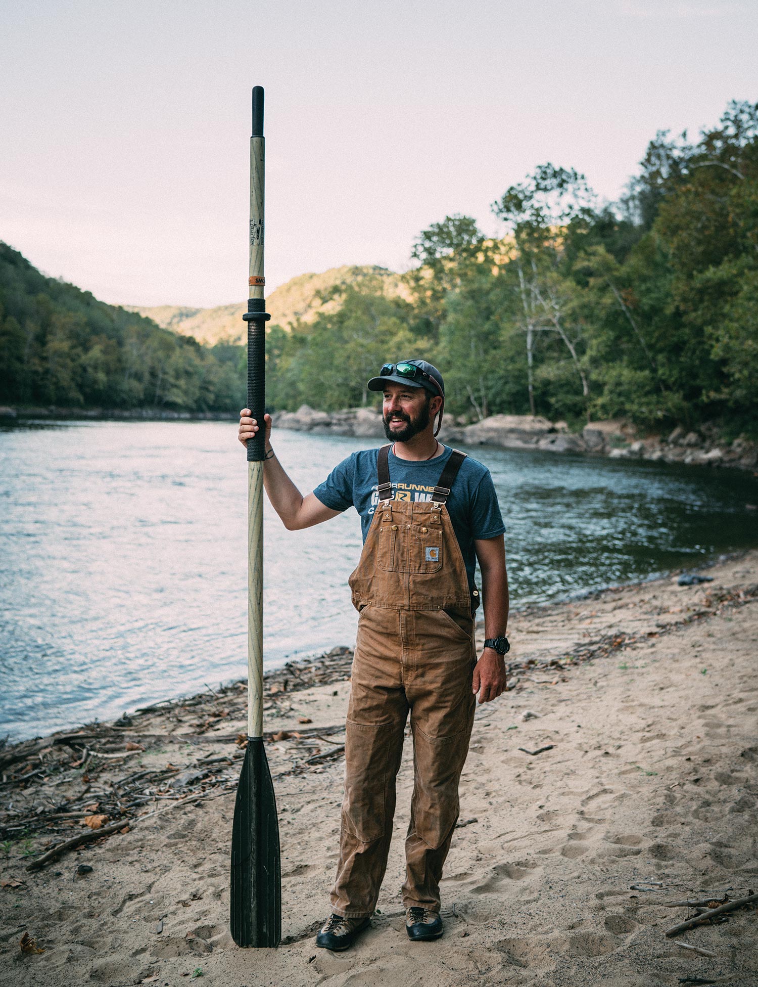 rafter wearing overalls stands on sandy riverbank and holds large boat paddle up vertically