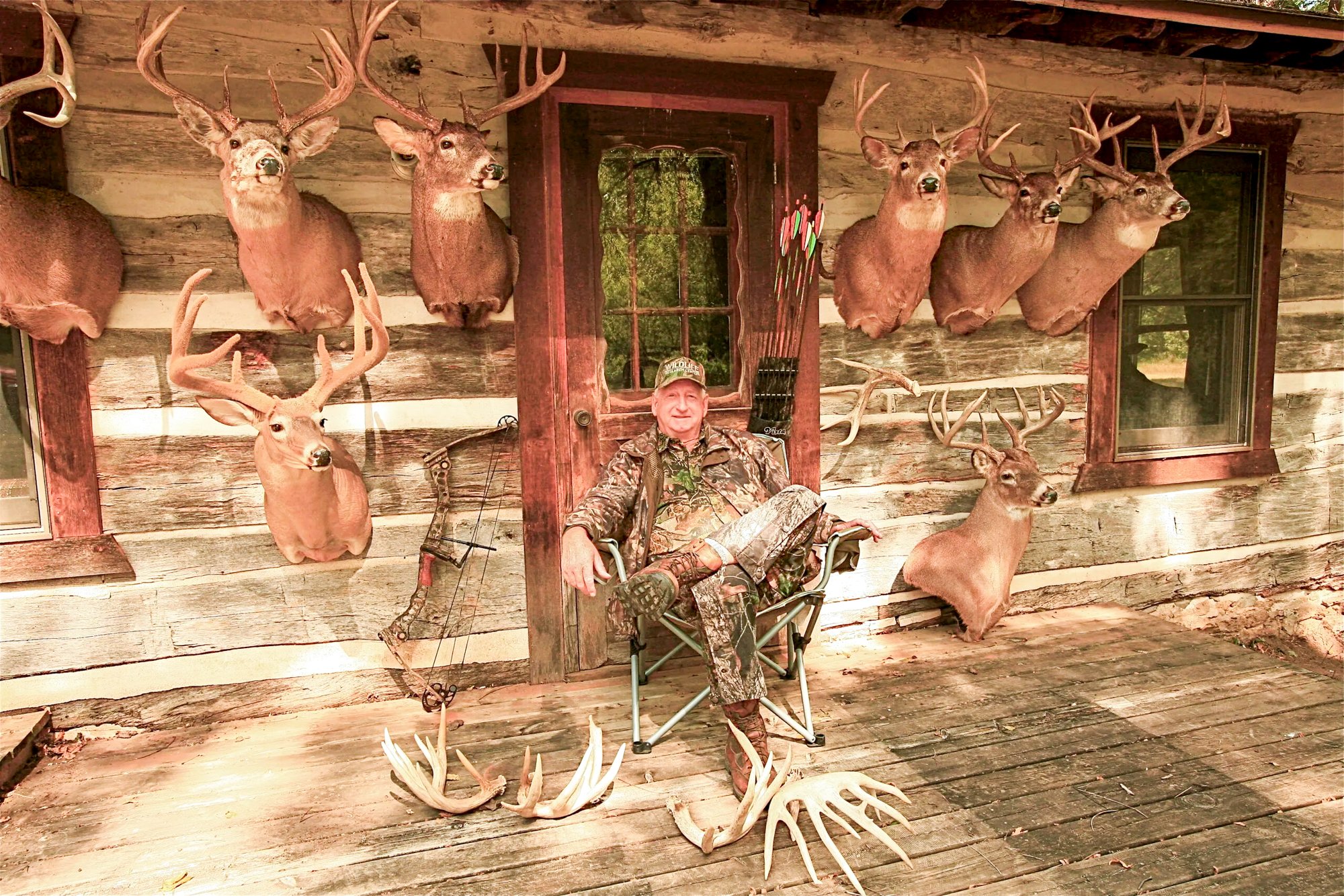 A hunter sits on a porch with a bunch of nice bucks on the wall.