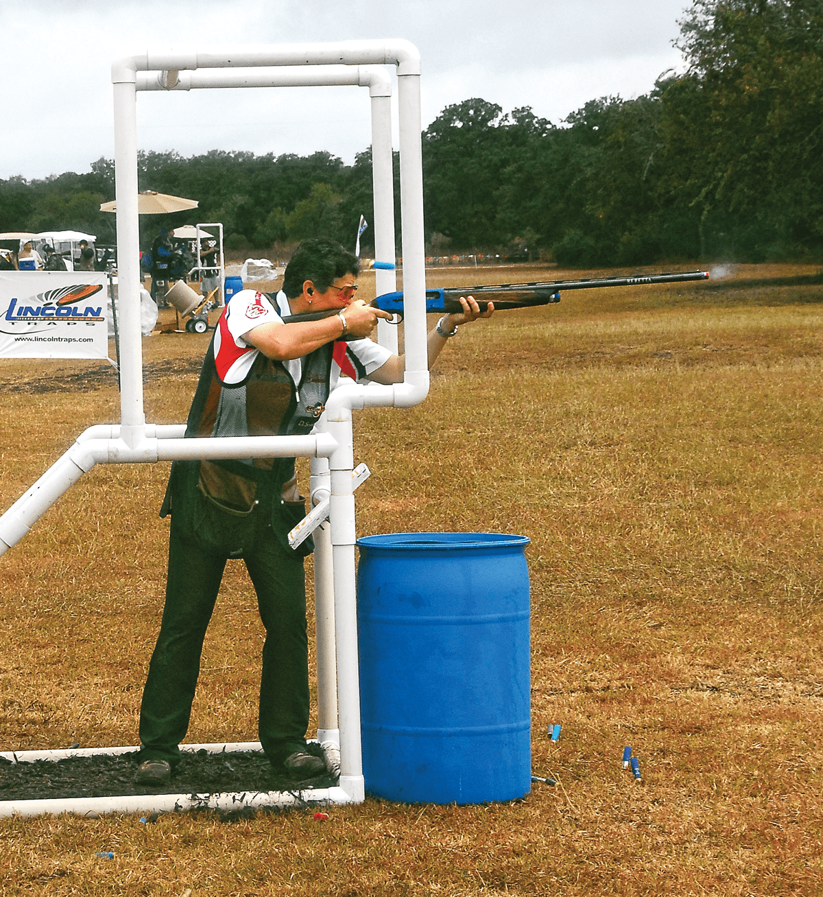 Woman shooting skeet at a pvc-pipe station, shooting vest, white shirt, blue range barrel