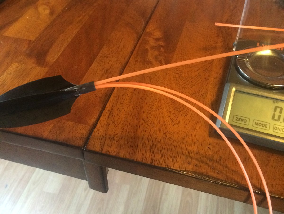 A broadhead with three strands of weed wacker line wedged in it, on a wooden table beside a scale.