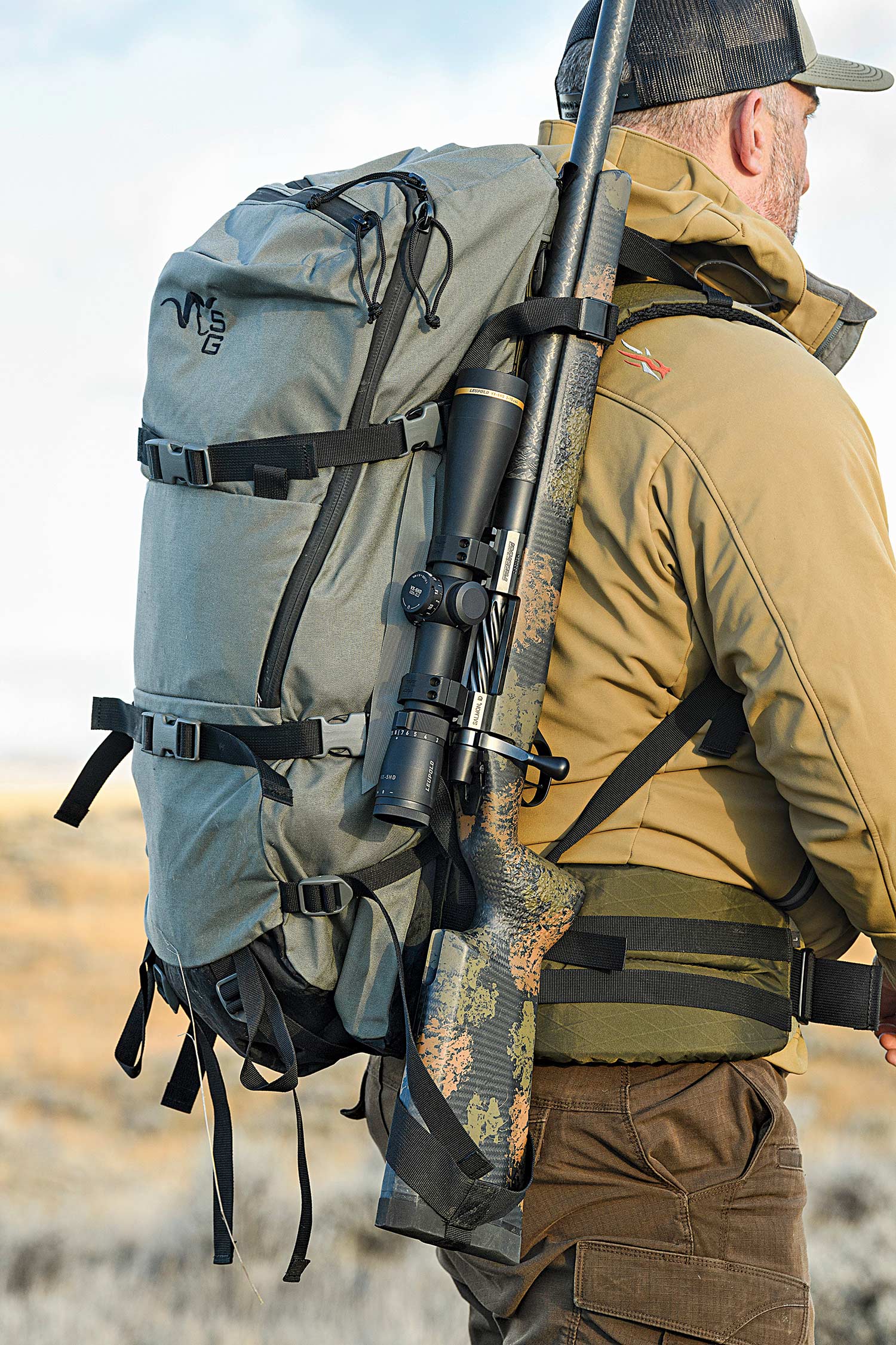 Hunter walking through a field with a backpack and rifle.