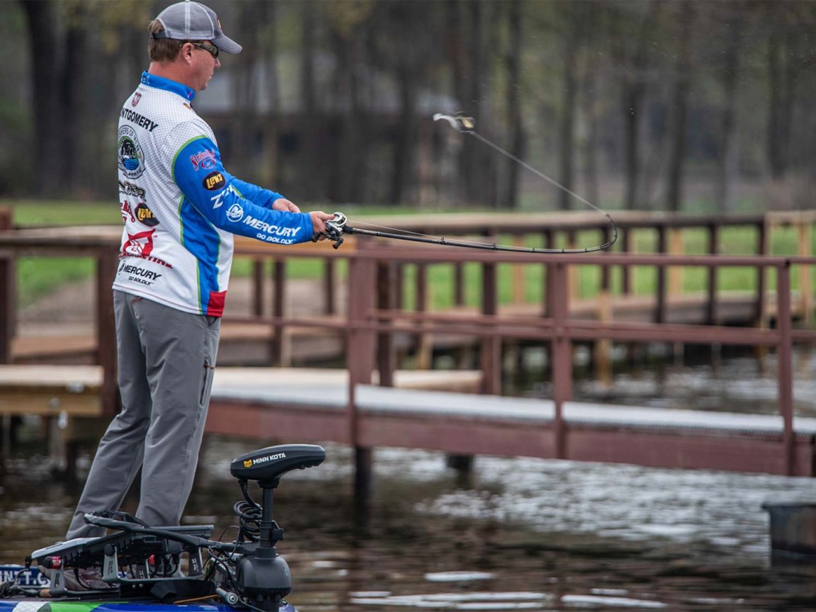 A male angler fishing off the front of a boat near a pier.