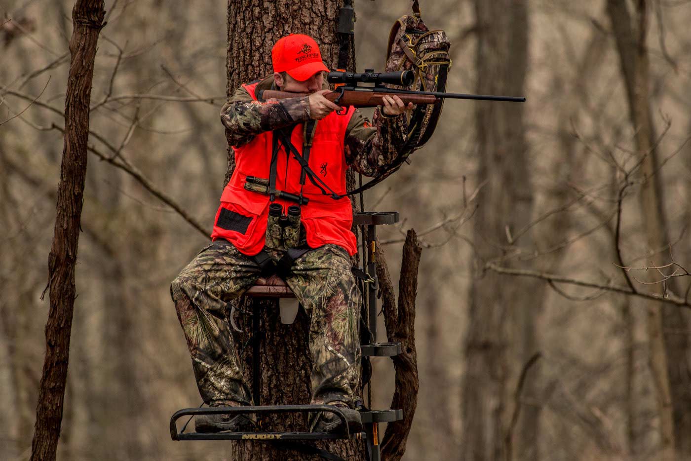 A hunter in an orange vest and hat fires a rifle from a treestand.