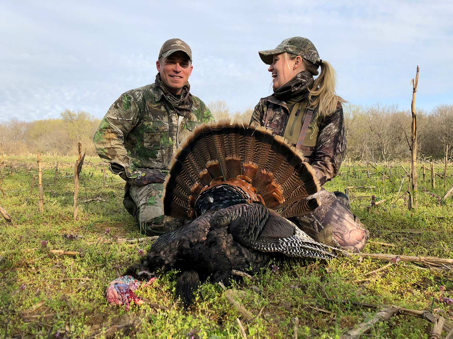 Two hunters kneeling behind a turkey in a field.