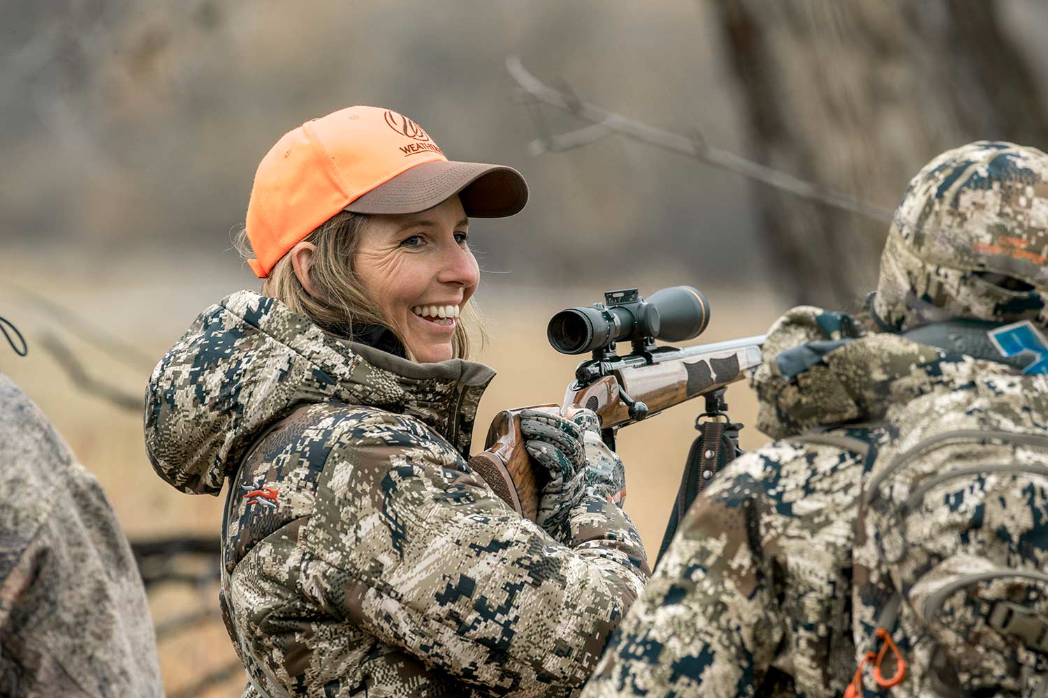 A female hunter smiles and aims a rifle with the help of hunting mentors and shooting sticks.