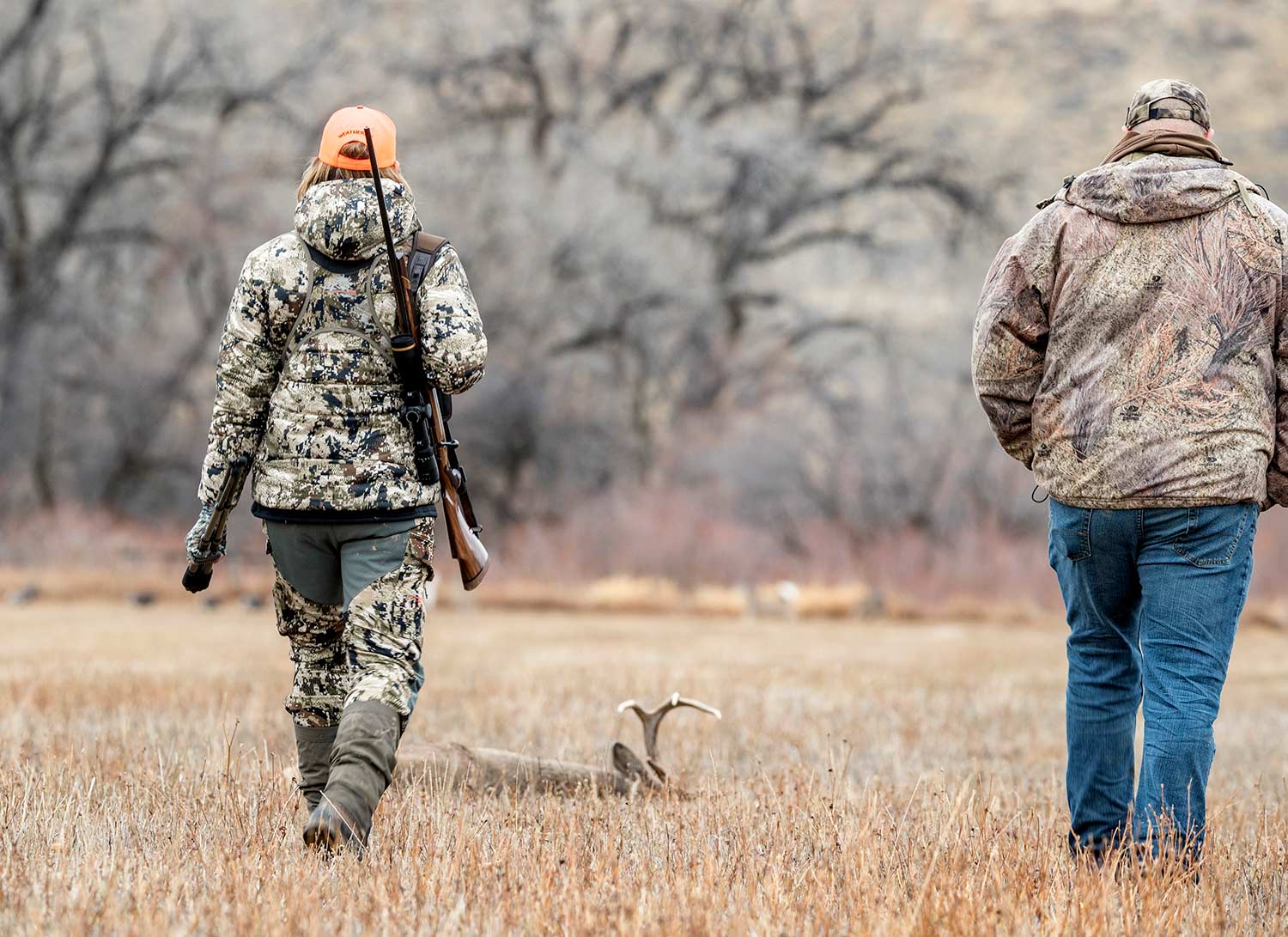 Two hunters decked out in gear walk through a field.