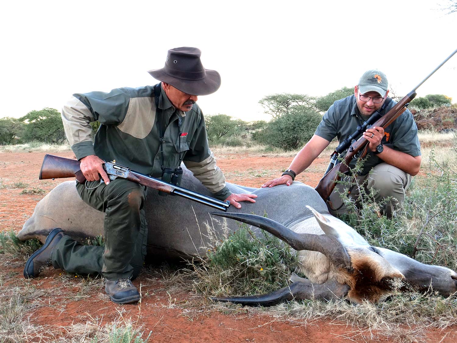 Two male hunters kneeling next to a downed pronghorn while holding rifles.