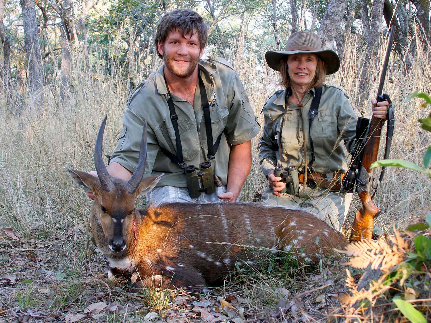 A male and female hunter kneels behind a bushbuck ram.