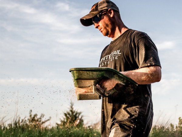 A man in a cap spreads grass and plant seed in a field.