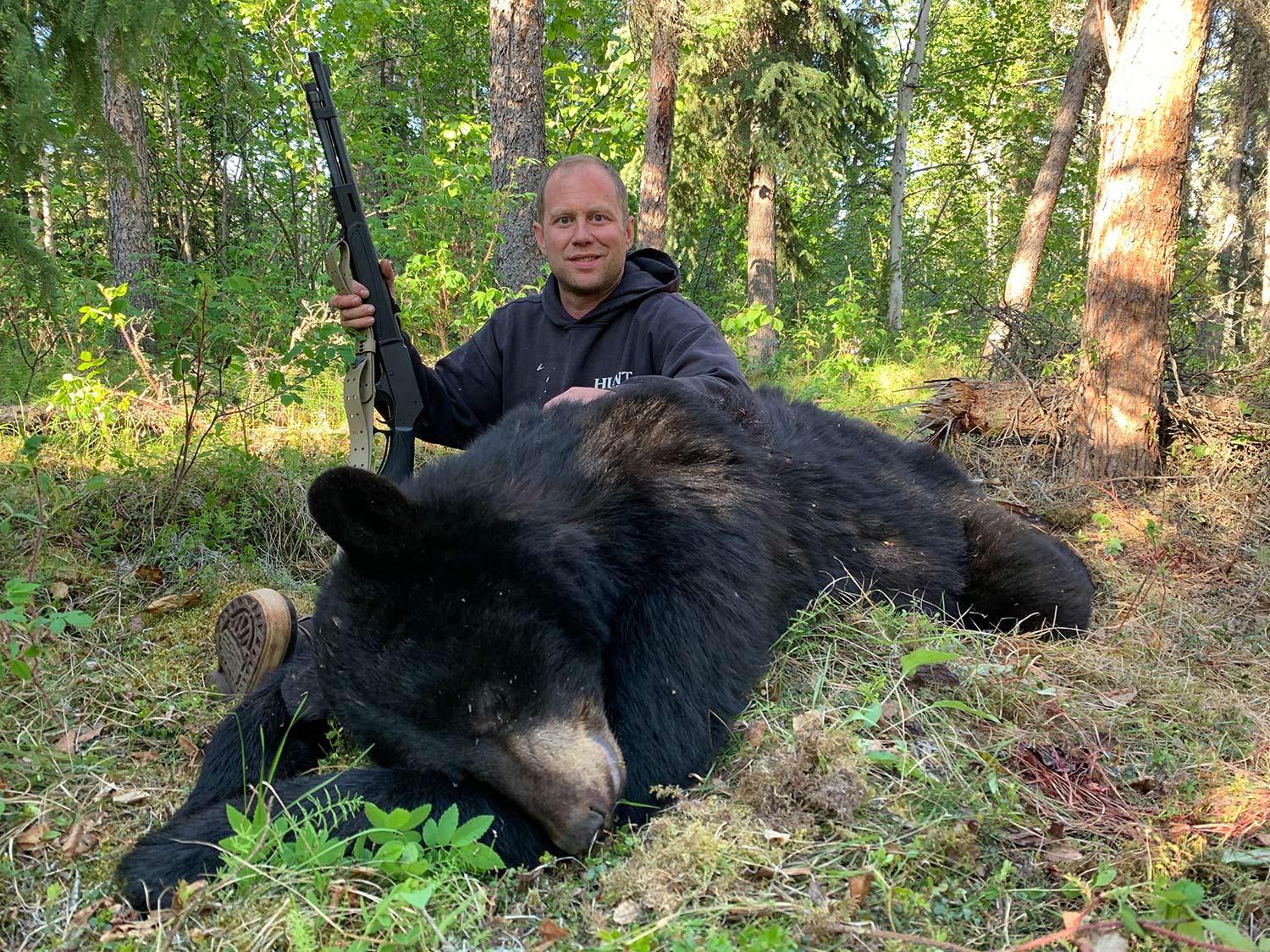 A hunter holds a rifle while kneeling behind an Alaskan Black Bear.