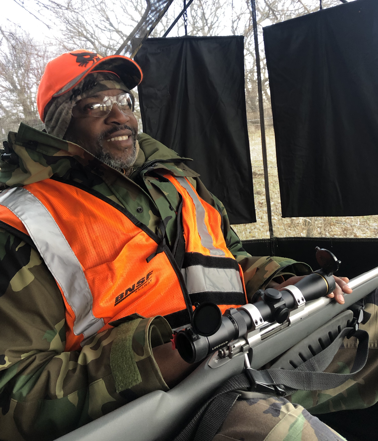 A hunter sitting inside a screened-in blind with camo, blaze orange, and a rifle across his lap.