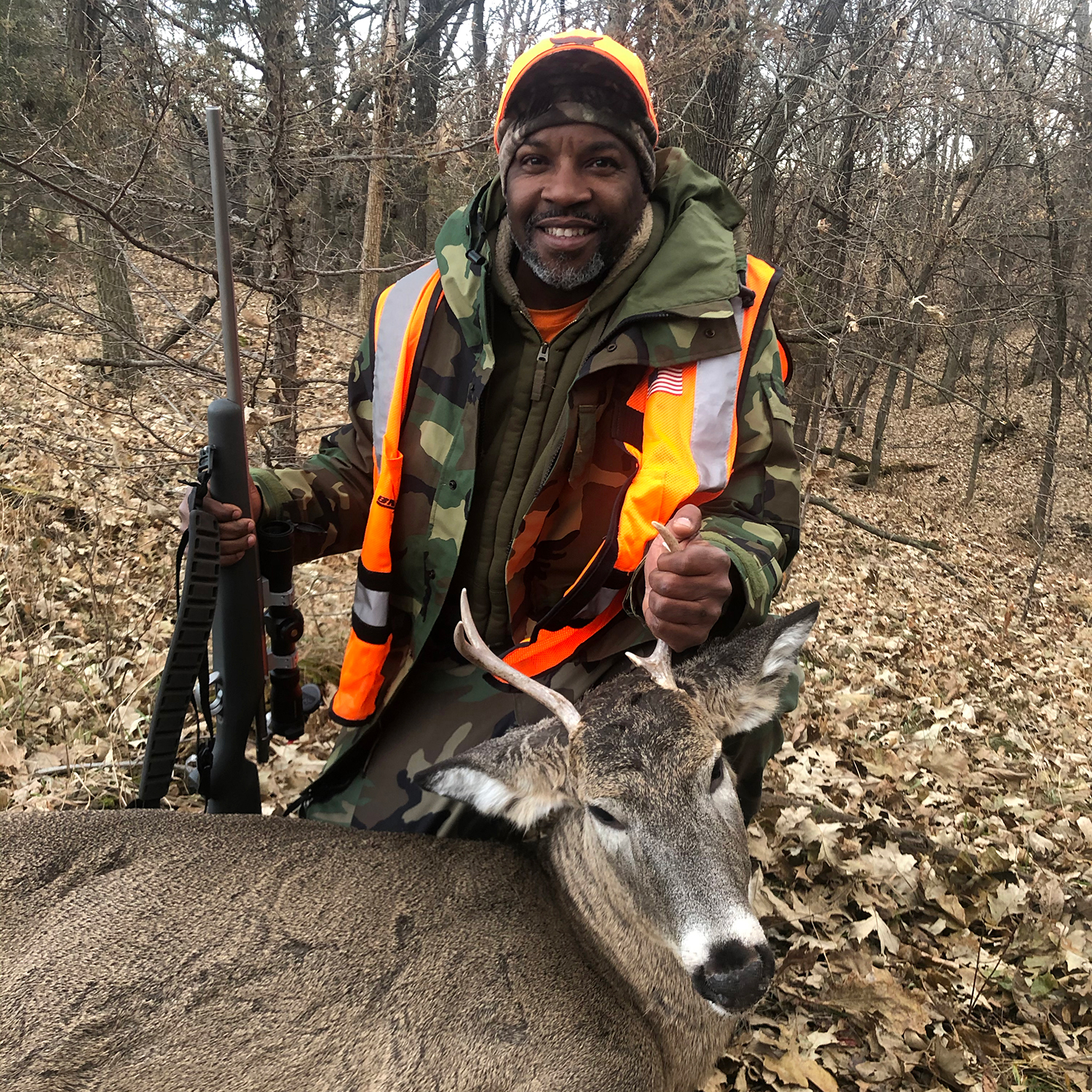 A black man in hunters camo and orange vest kneels behind a deer in the woods.