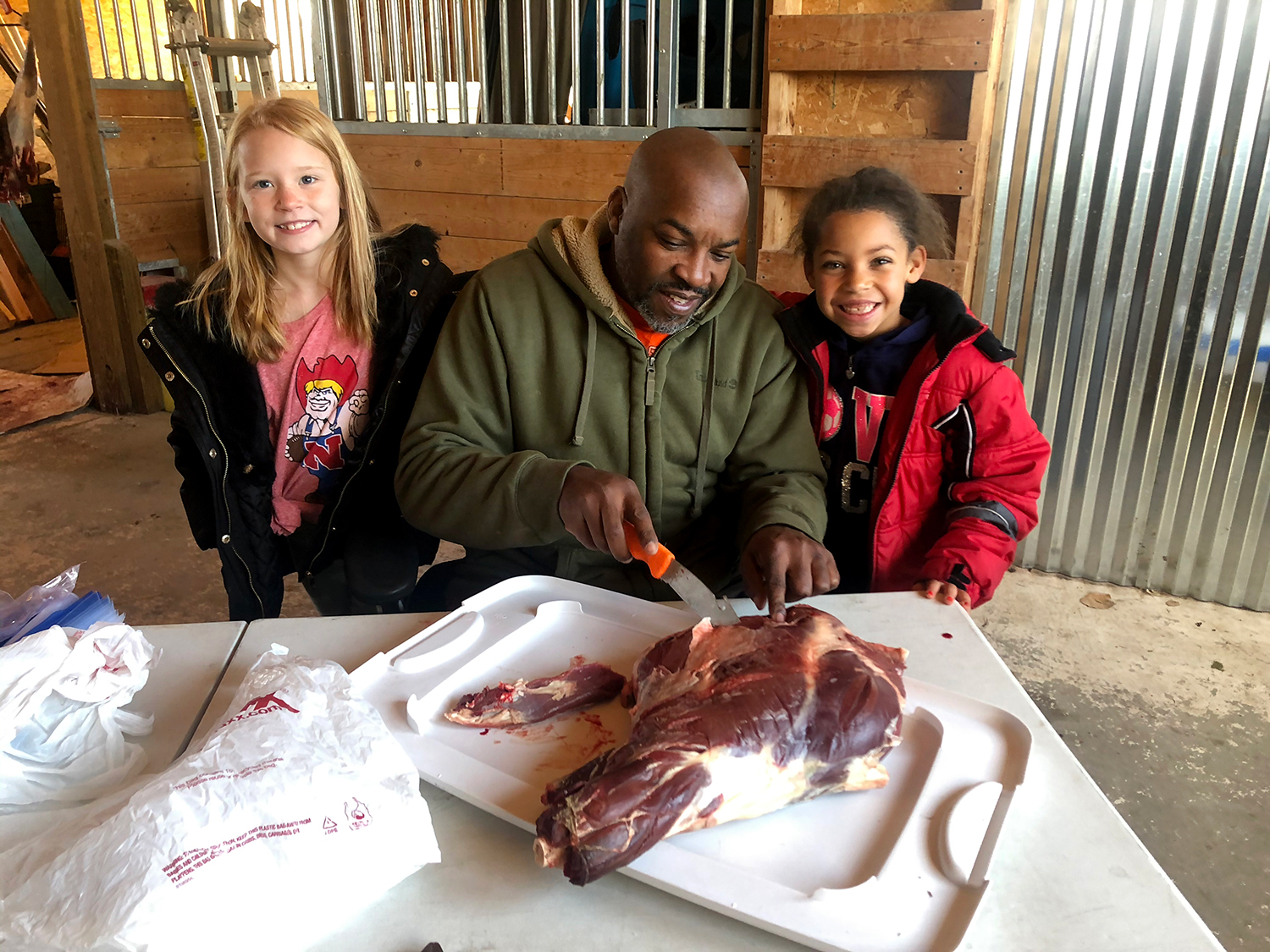 A man carves and butchers deer meat while two kids stand beside him.