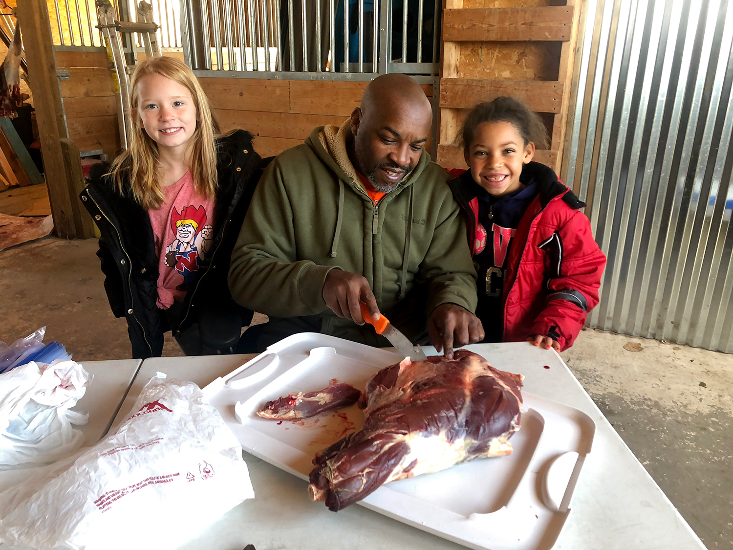 A black man carves and butchers deer meat while two kids stand beside him.