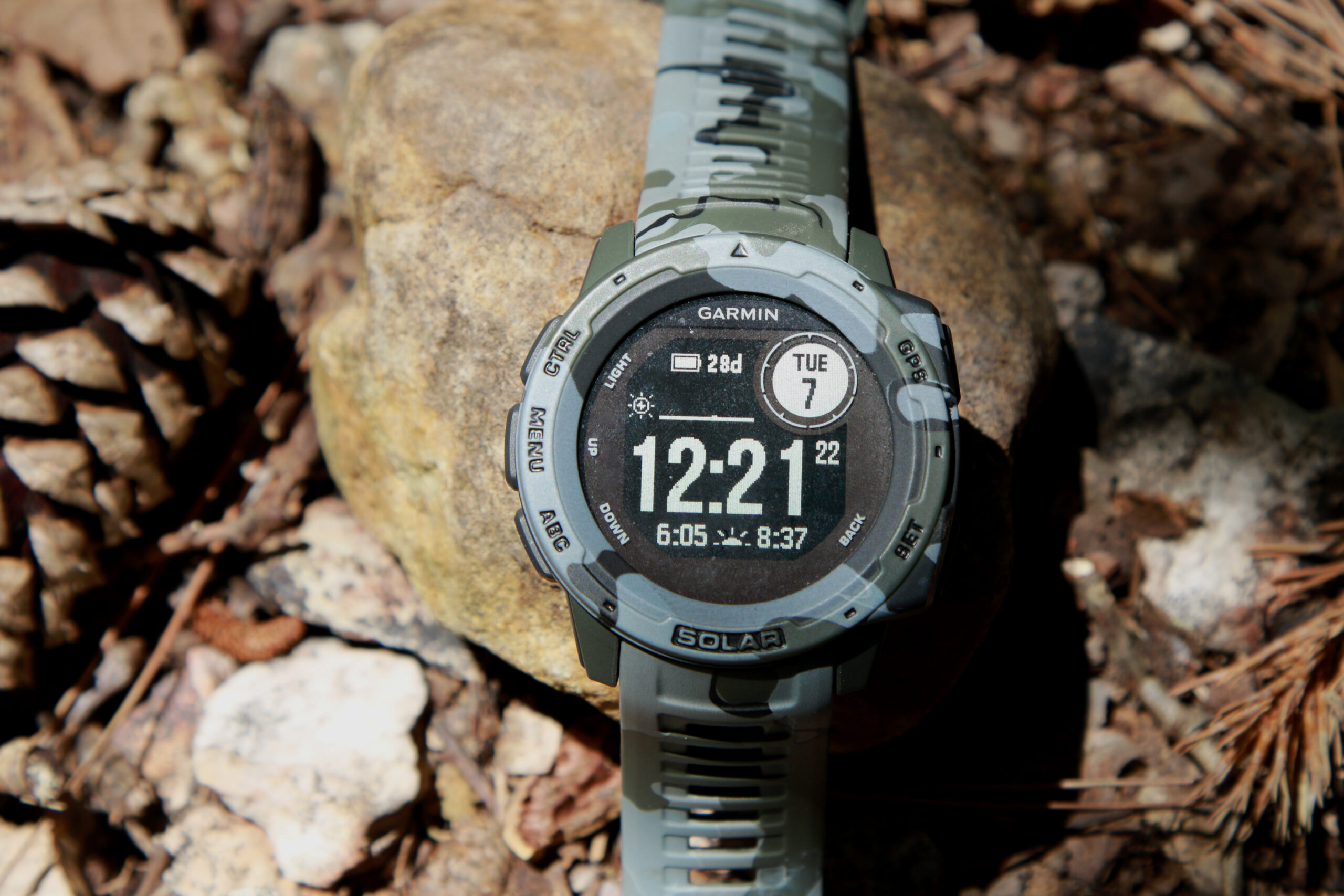 A camo watch displaying the time 12:21 and a battery life of 28 days on a rocky background.
