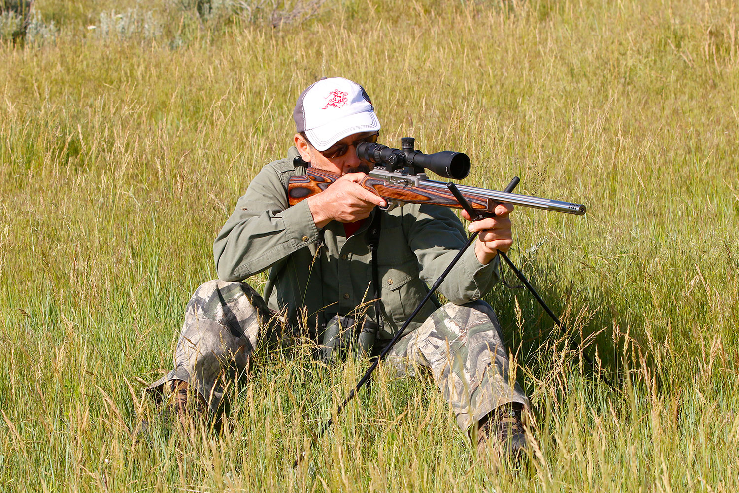 A man sitting in tall grass while aiming a rifle using a shooting stick.