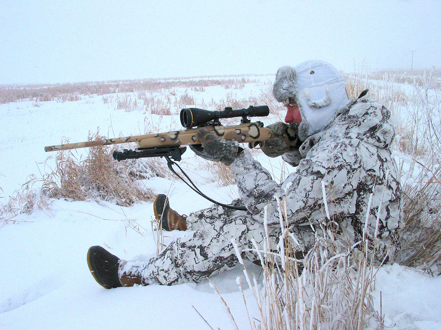 A man sitting in the snow and aiming a rifle.
