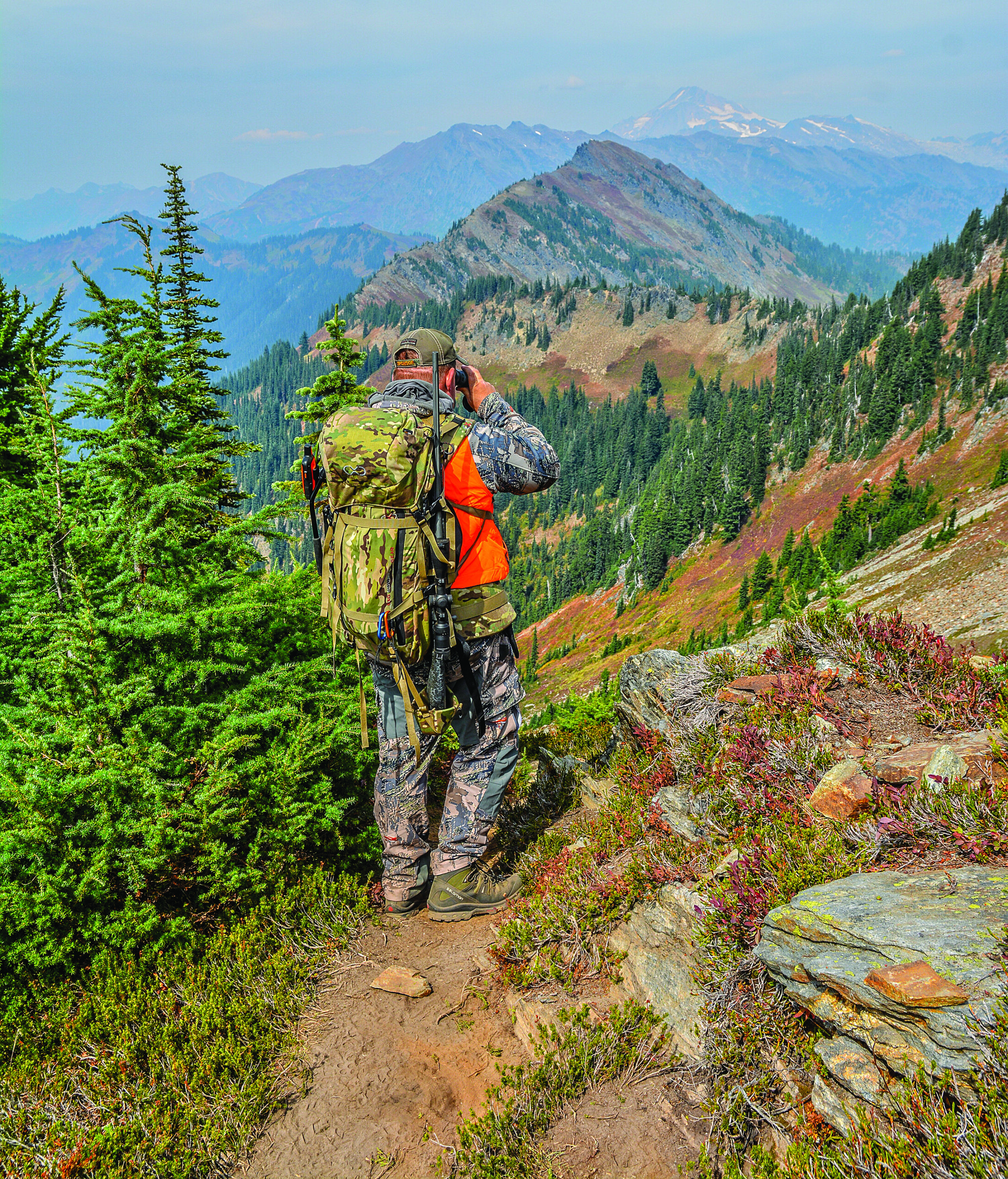 A hunter in blaze orange with a pack glassing in the mountains.