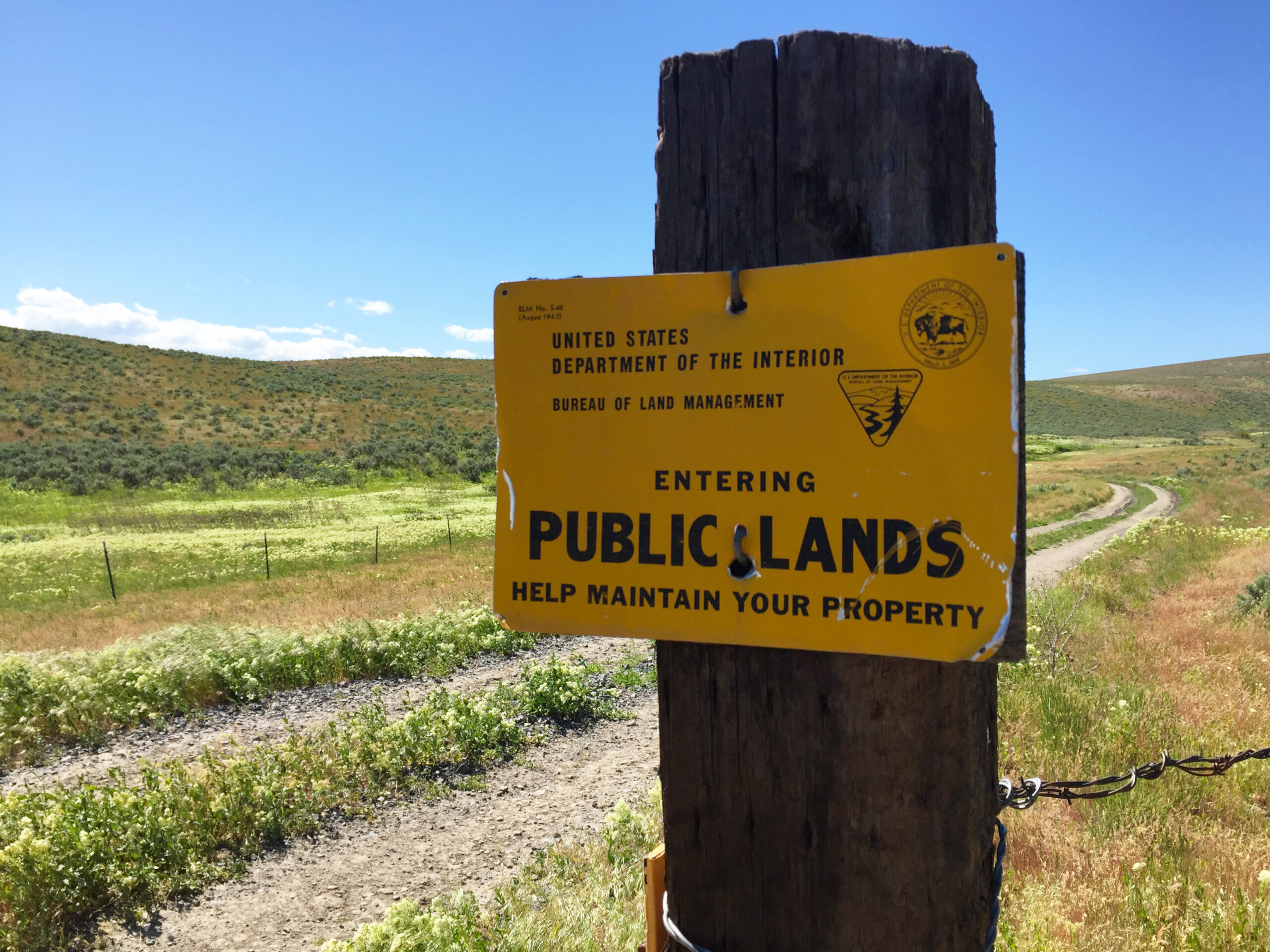 A yellow sign that reads "Entering Public Lands" on a weathered fence post in front of a two-track road