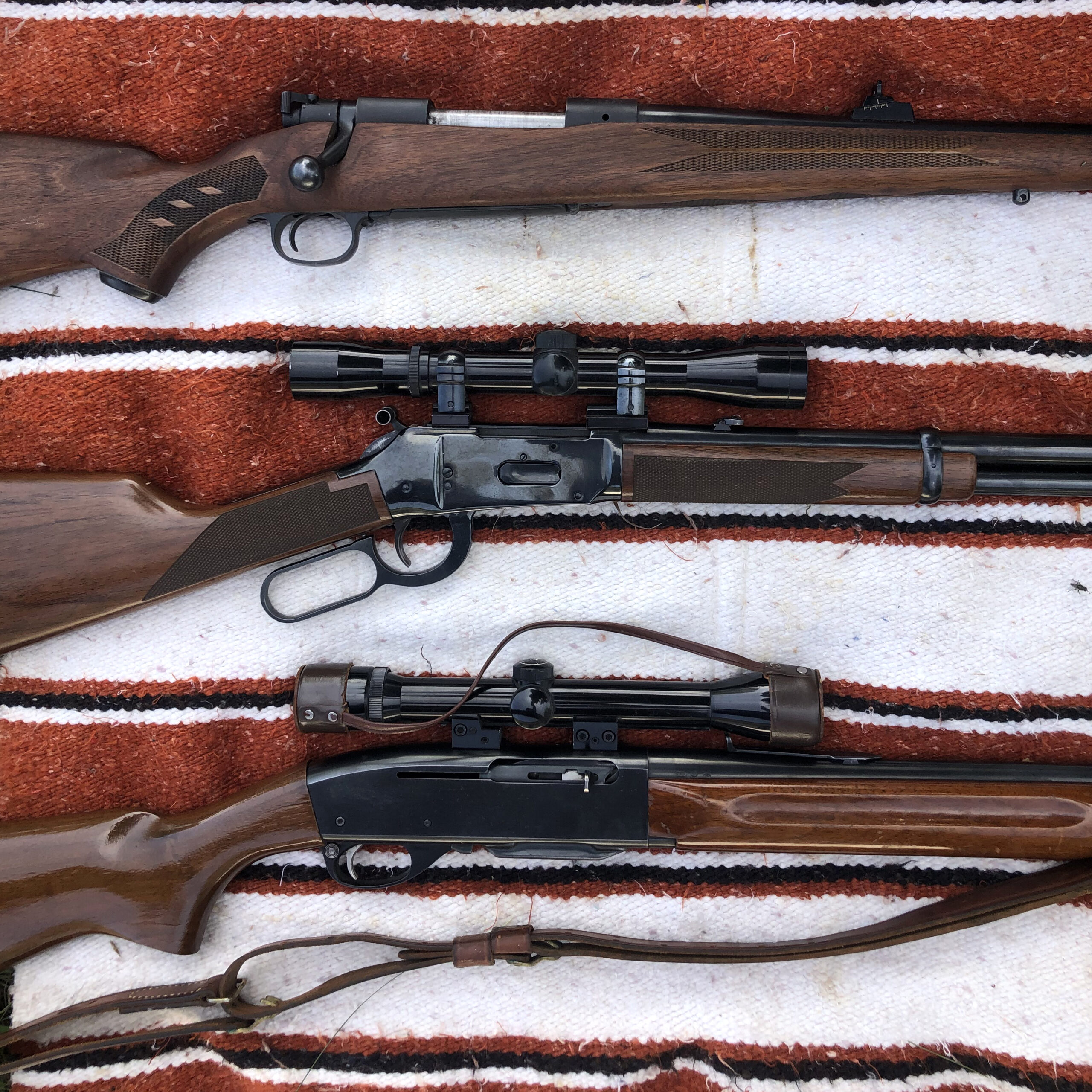 Three old rifles, two with scopes, on a woven red, white, and black blanket.