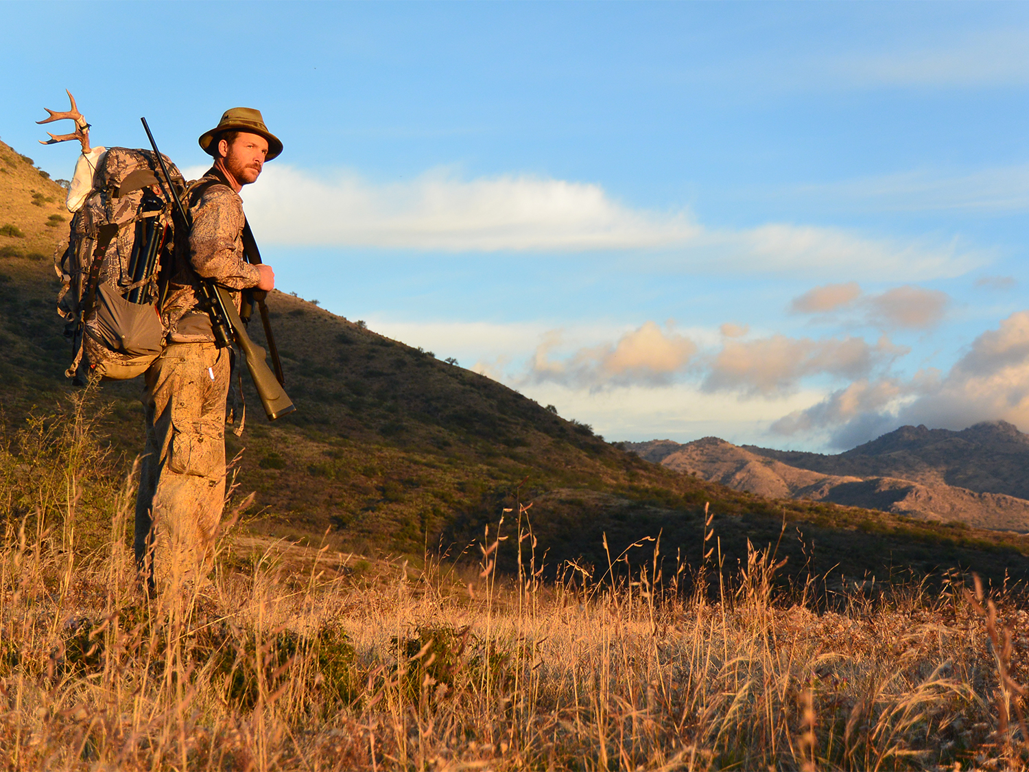 A man with a rifle and backpack in a large hillside.