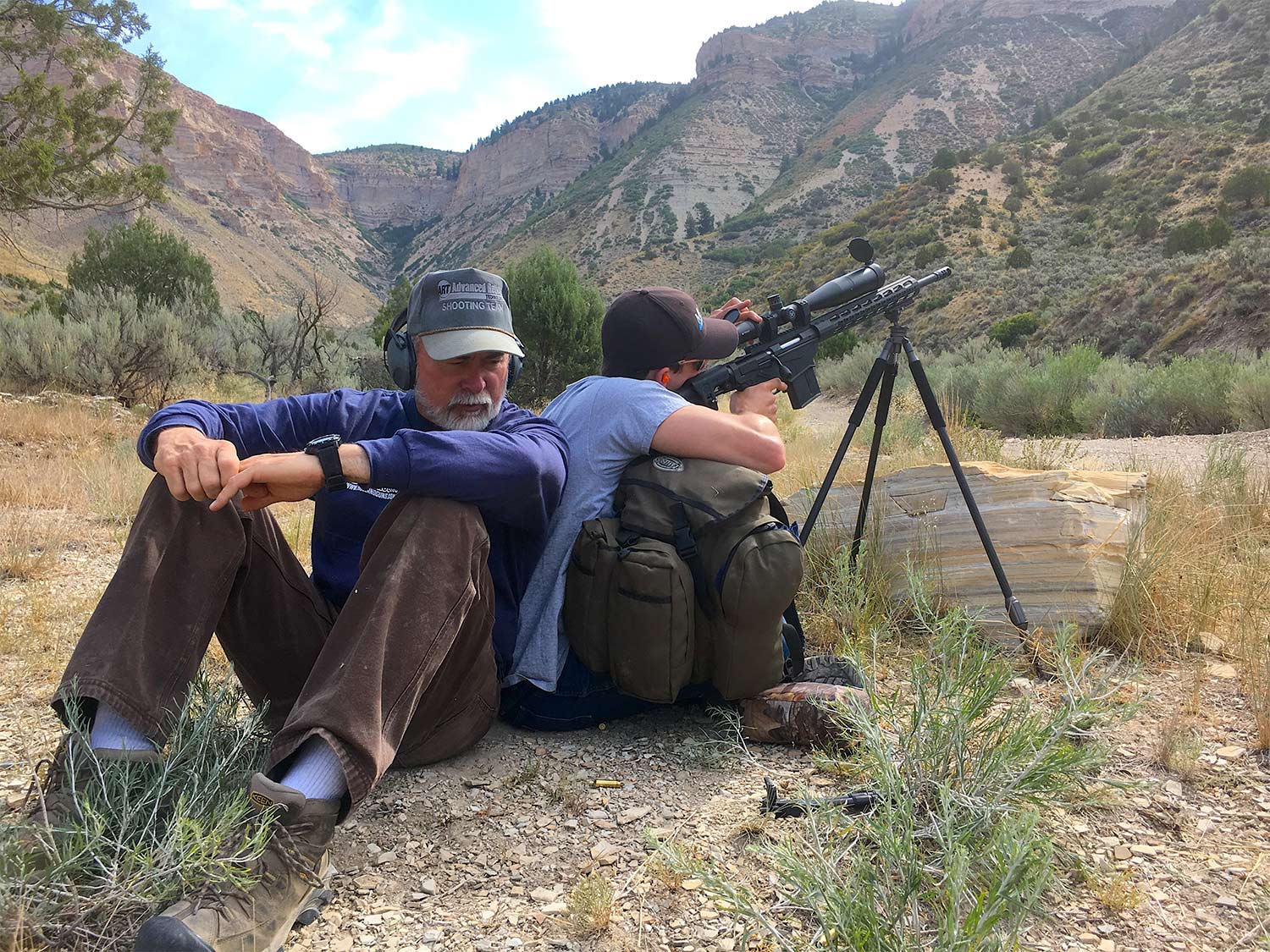 Two men sit back-to-back while one aims a rifle.