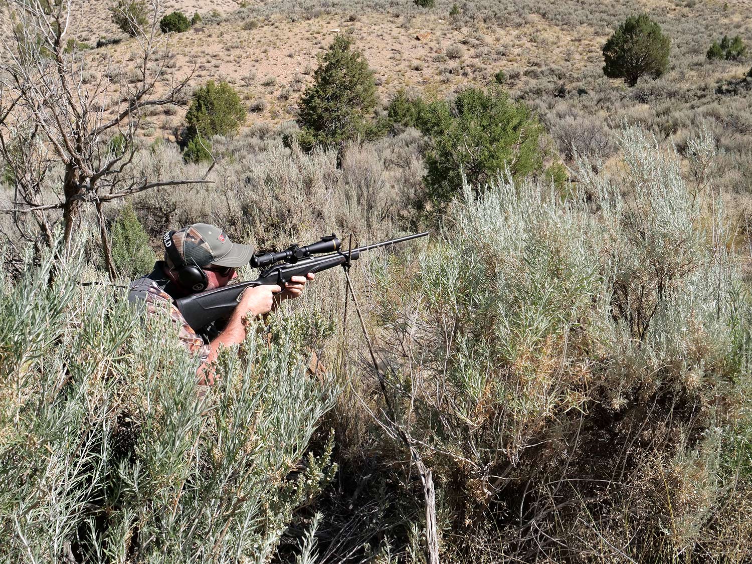 A concealed hunter inn camo aims a rifle in a field.