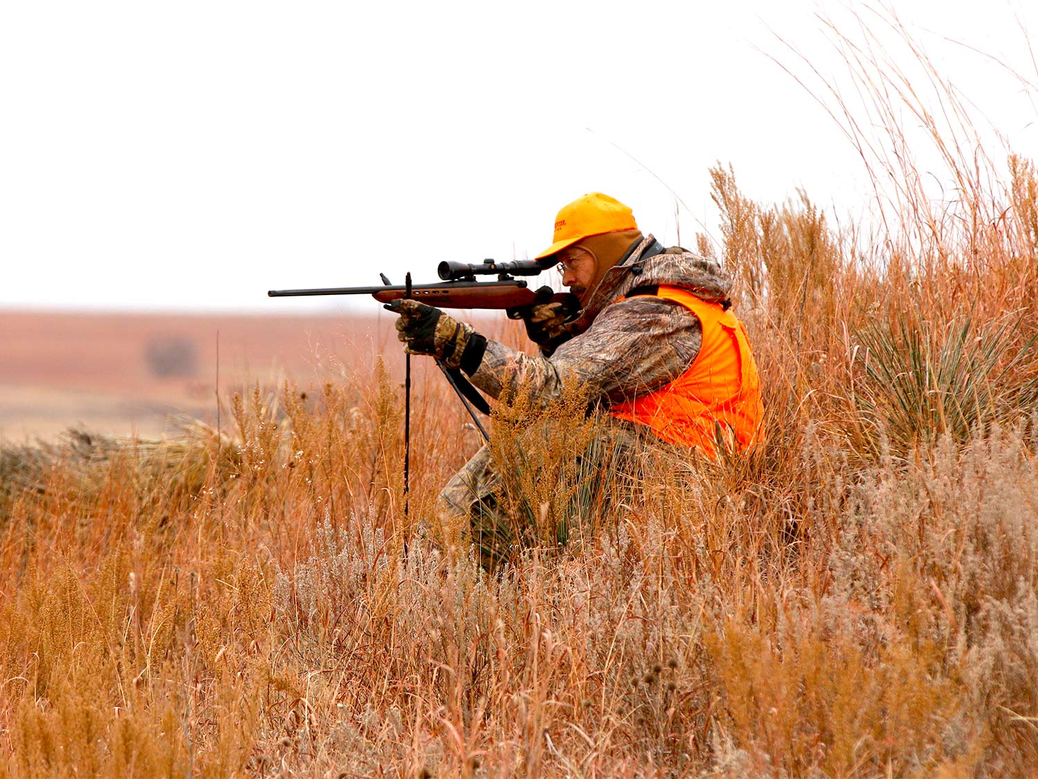 A hunter in camo and orange aims a rifle in a field.
