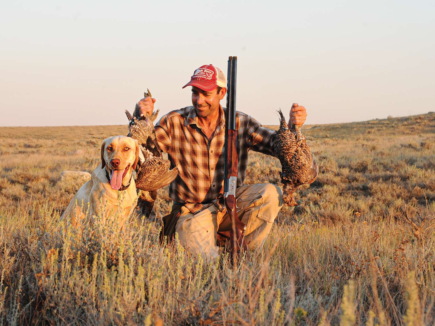 A hunter holds up ruffed grouse next to a hunting dog.