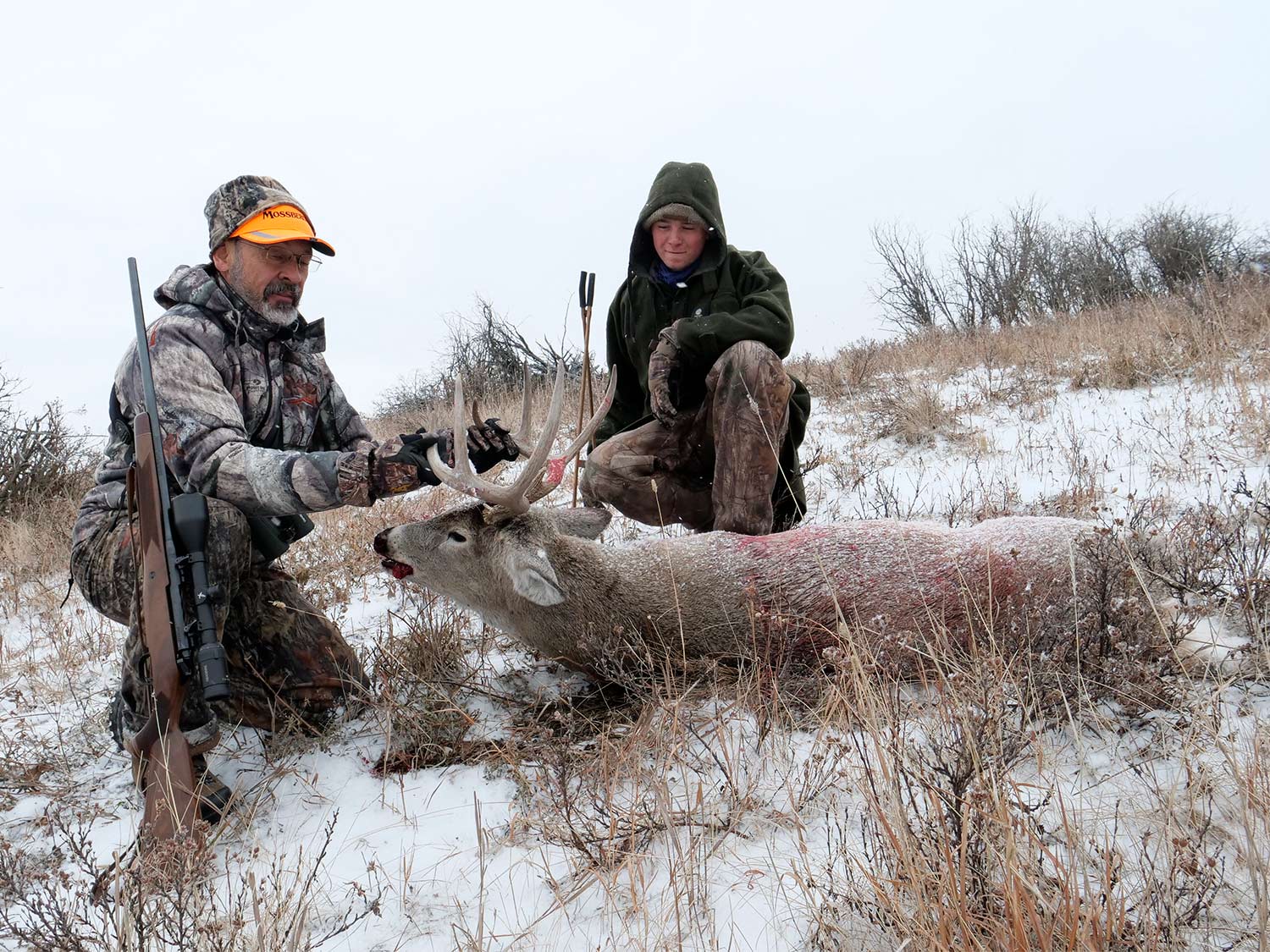 Two hunters kneeling behind a whitetail buck in the snow.
